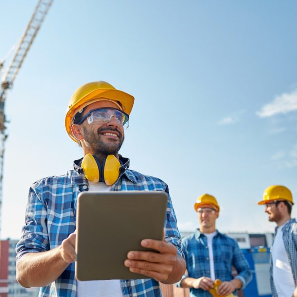 Smiling Construction Worker With A Hard Hat And Safety Glasses Holding A Tablet At A Construction Site, With A Crane In The Background And Colleagues Discussing Plans, Highlighting The Satisfaction Team Members Can Experience When Being In Sync With Eachother And Their Projects With Sharepoint | Hingepoint